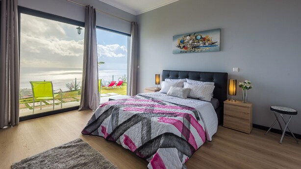 A bedroom with a large bed, a view of the ocean, a painting on the wall, and a small table with a lamp. - Gorgeous, Modern Home with Breathtaking Views (Arco da Calheta)