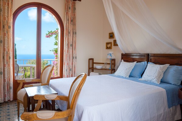 Ein Schlafzimmer mit einem großen Bett, zwei Sesseln, einem kleinen Tisch und Meerblick durch ein Fenster.