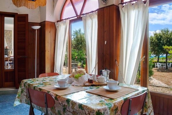 A dining area with a table set for two, featuring a floral tablecloth, white plates, and a metal pitcher. The room has wooden walls and a large window with white curtains.