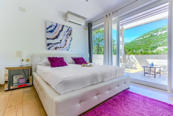 A modern bedroom with a large bed, a view of a green landscape, and a balcony with outdoor furniture. - Villa Oasis, 5 bedrooms, heated Pool, Jacuzzi, BeachFront by MyWaycation (Smokvina)