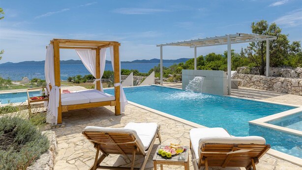 Un área junto a la piscina con una pérgola de madera, sillones de descanso y una mesa con frutas y bebidas.