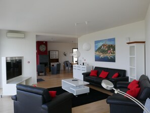A modern living room with a black sofa, a white coffee table, and a flat-screen TV. There is a dining area with a red wall and a clock. A painting of a coastal town is on the wall. - Villa Paradise with pool and sea views, beautiful, 7 people (Vrbnik)