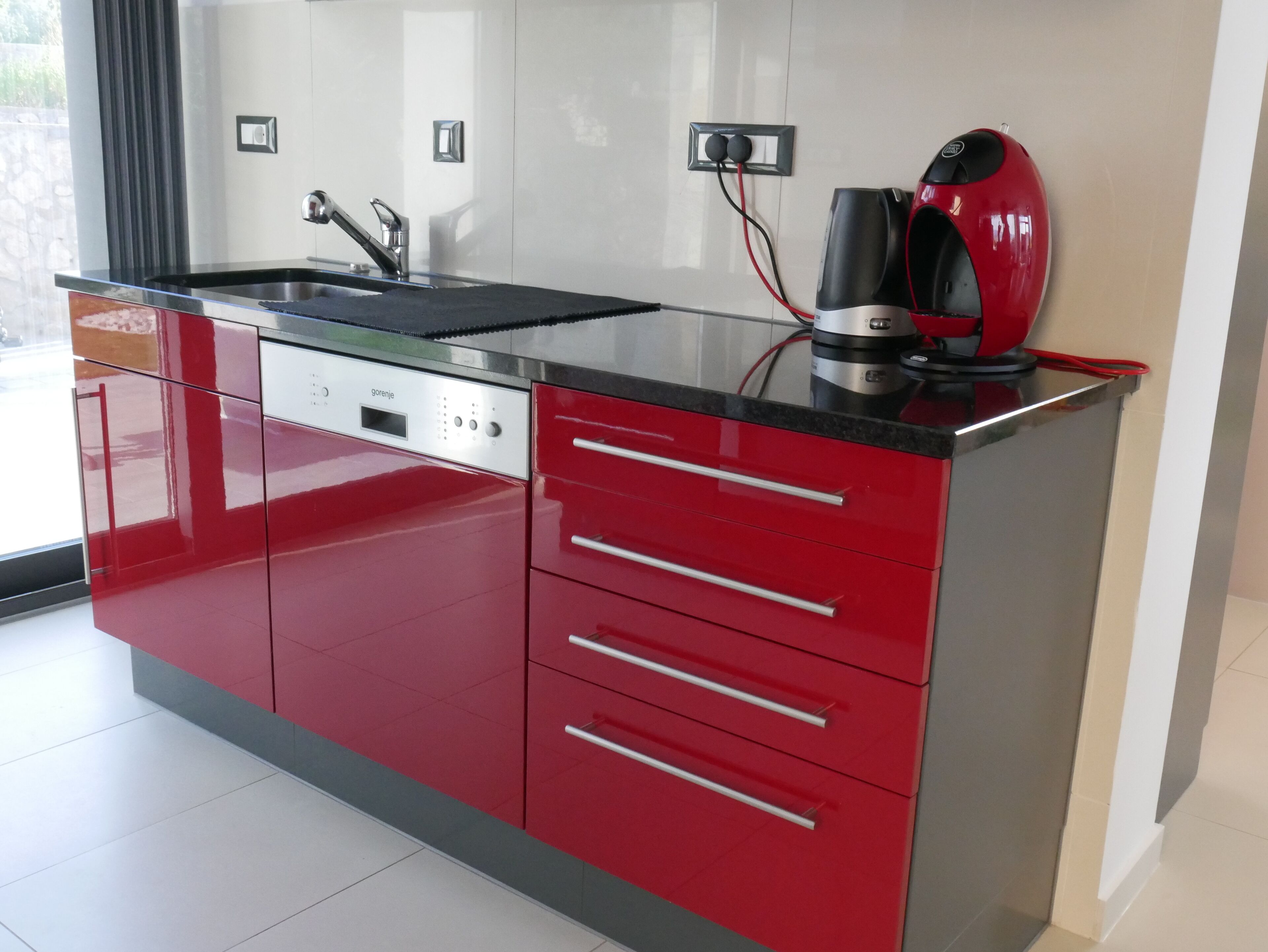 A modern kitchen with red cabinetry, a stainless steel sink, and a red kettle.
