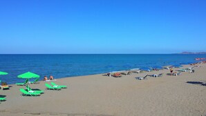 A beach with rows of green sun loungers and blue umbrellas, a clear blue sky, and calm sea.