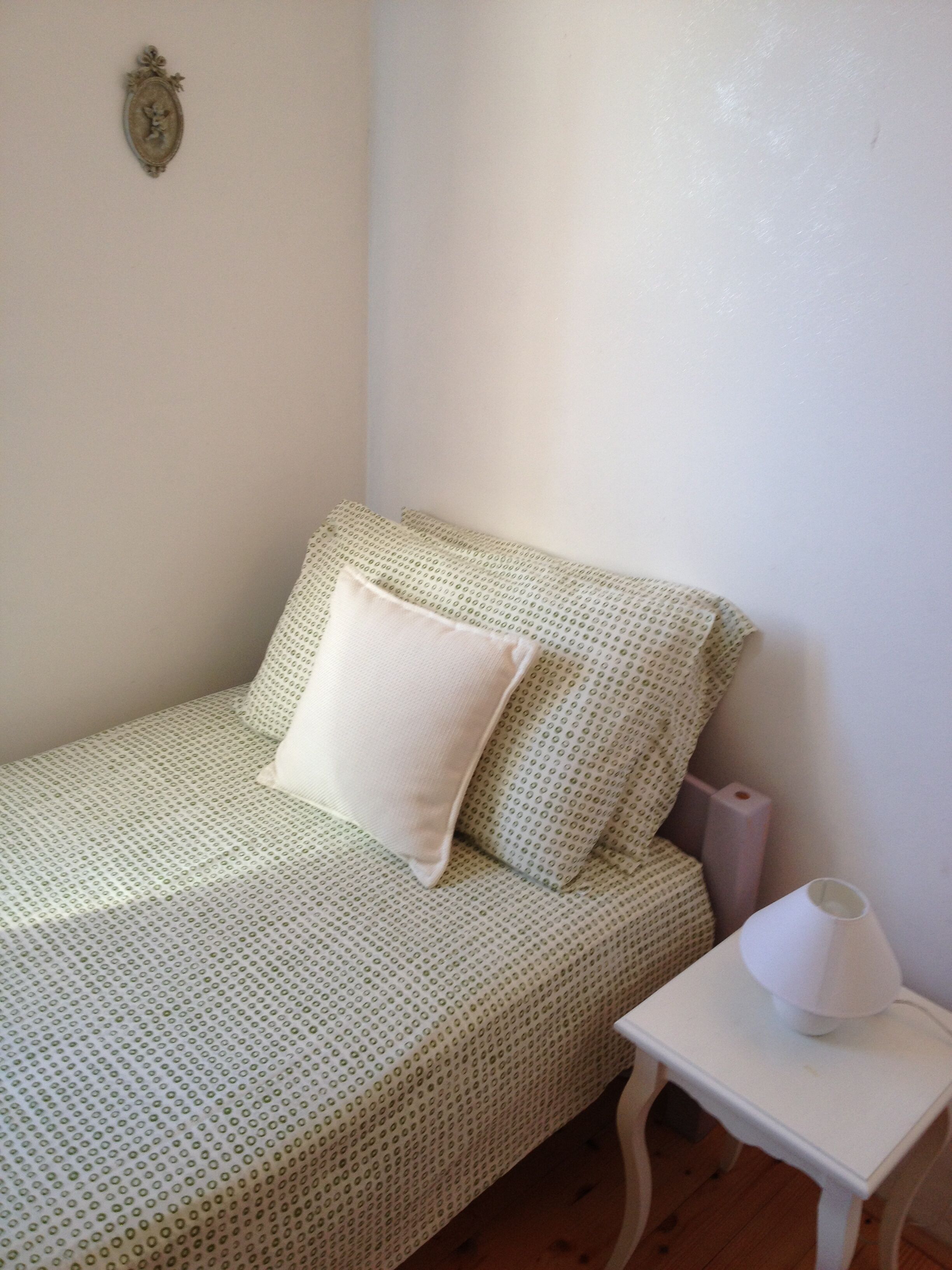 A small, single-bed room with a patterned bedspread, a white pillow, a bedside table with a lamp, and a wall-mounted decorative item.