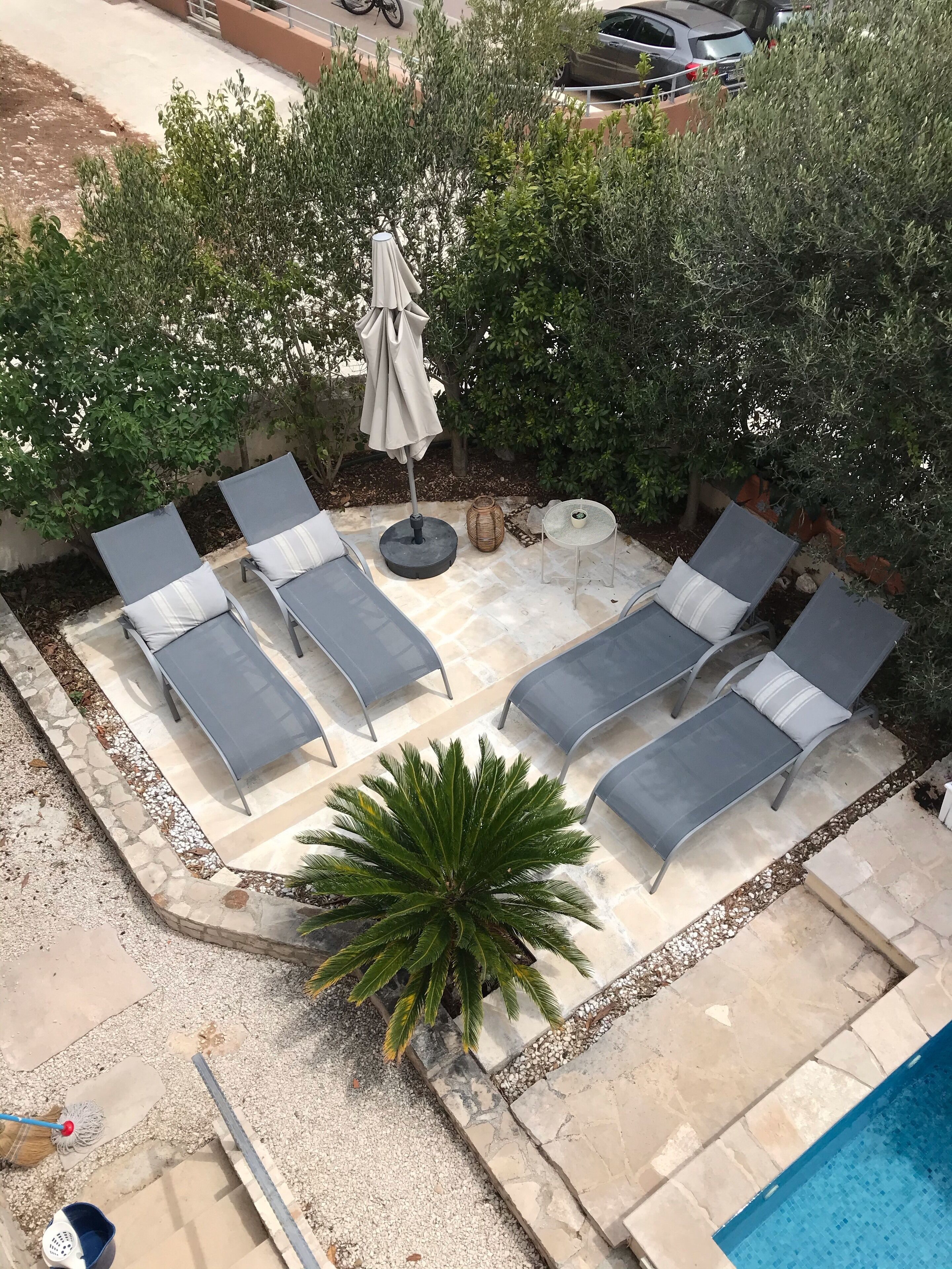 An outdoor seating area with lounge chairs and a table, surrounded by greenery and a swimming pool.