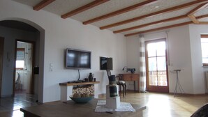 A spacious living room with wooden beams on the ceiling, a television, a sewing machine, and a large window. - Modern spacious apartment in a quiet south-facing slope. (Kandern)