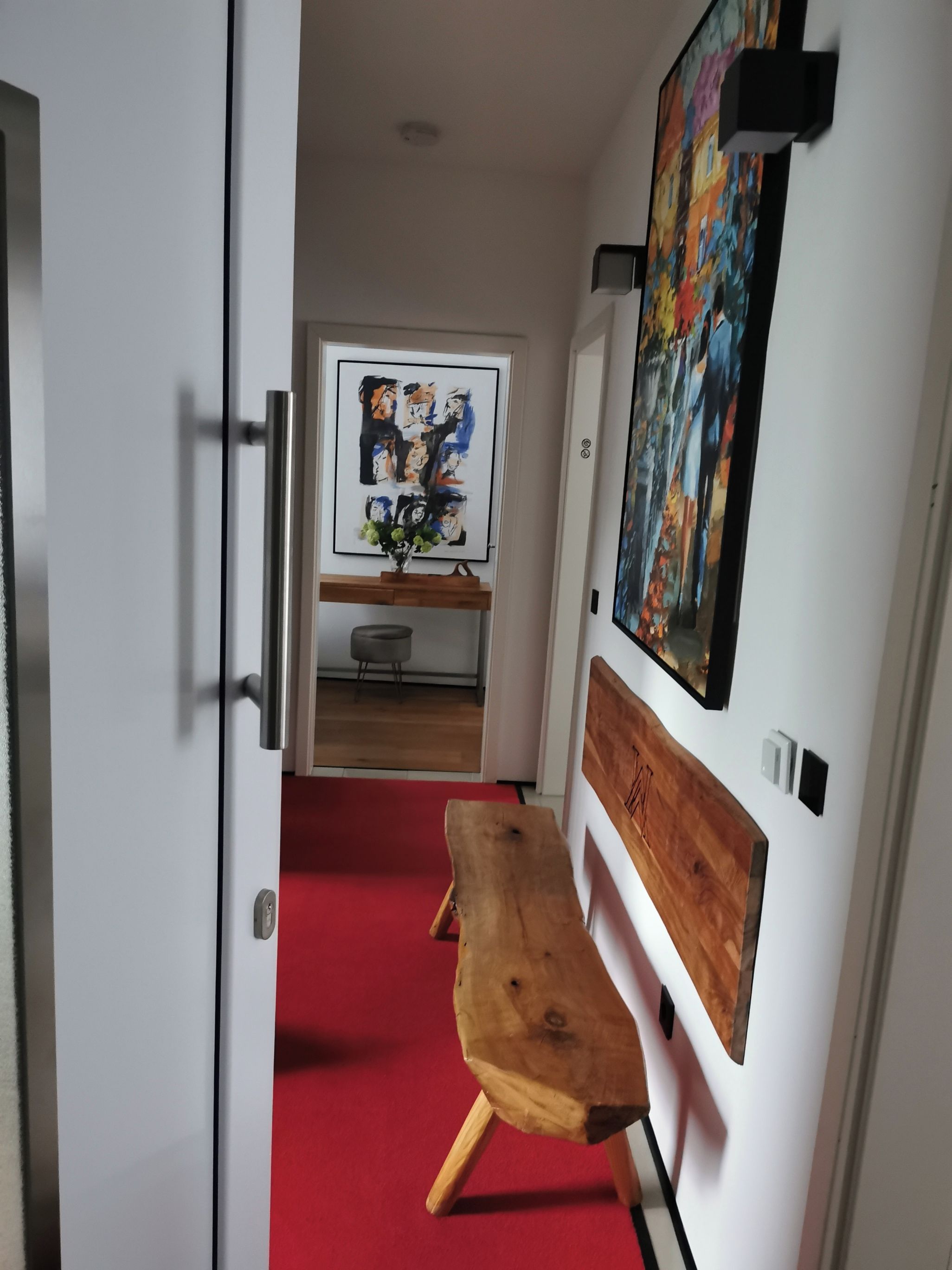 A hallway with a wooden bench, a red carpet, a door, and a wall with a painting.