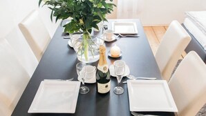 Une table dressée pour un dîner à deux, avec de l’assiette blanche, des couverts et des verres, une bouteille de champagne et une plante en pot.