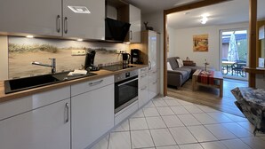 Una cocina moderna con armarios blancos, horno empotrado y cafetera. Se ve un comedor con mesa y sillas a través de una puerta abierta.