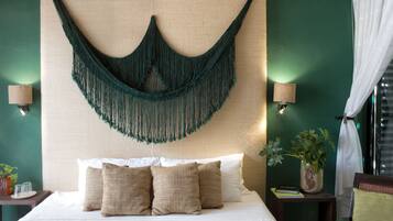 A bed with white linens and beige pillows, a green macrame wall hanging, and a potted plant on a nightstand.