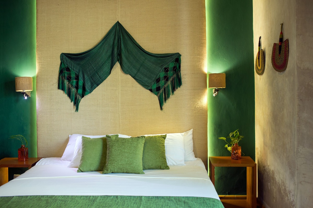 A neatly made bed with green and white bedding, a decorative green headscarf on the wall, and a small table with a vase of flowers.