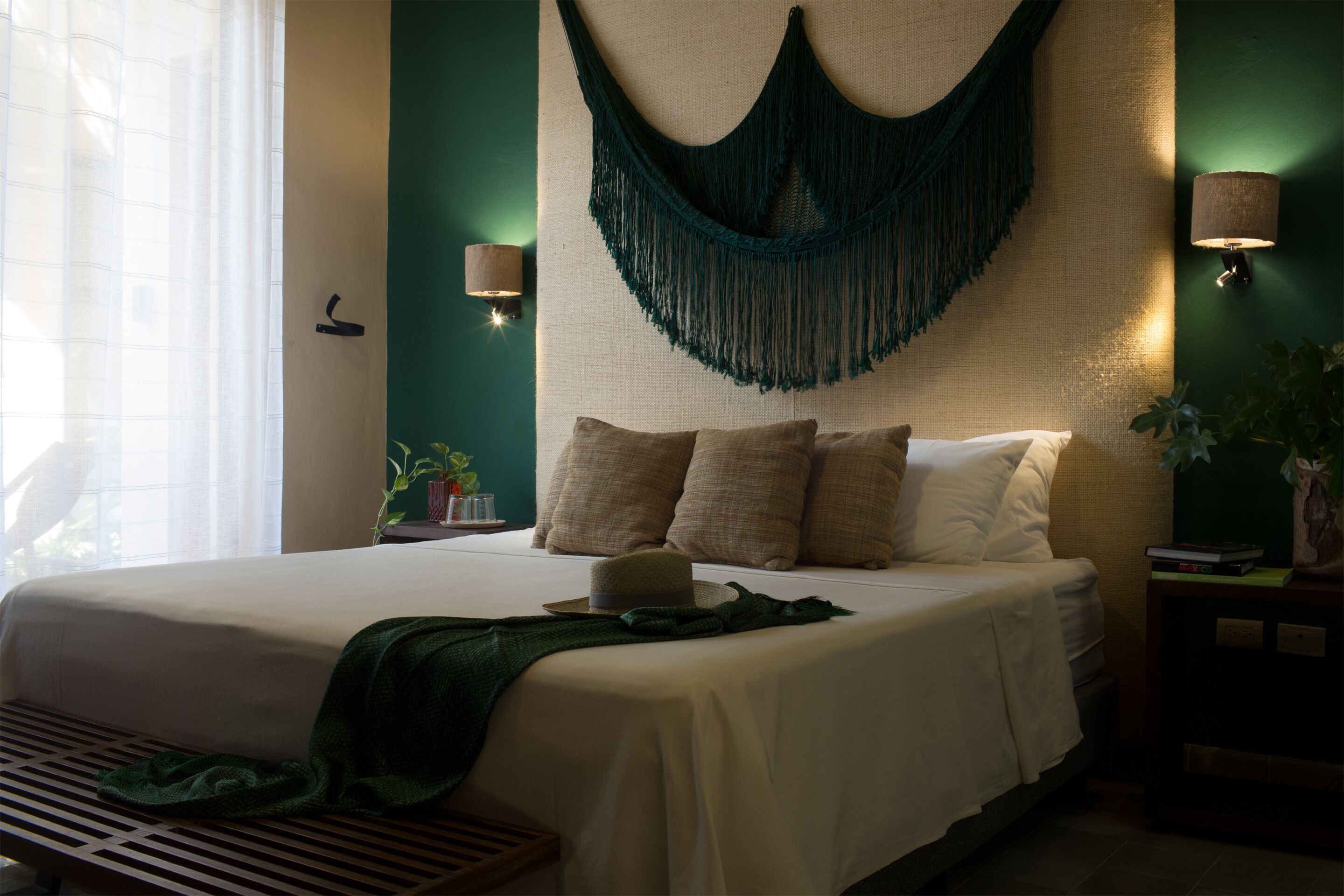 Un lit bien fait, agrémenté d’un plaid vert, d’une lampe de chevet et d’une décoration murale en macramé.