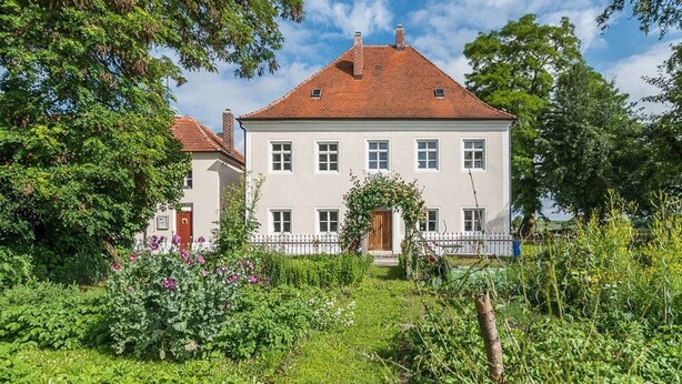 A two-story house with a red-tiled roof, surrounded by lush greenery and a white fence. - 3 Stars Holiday House - Historic ambience of the 18th century. Century (Niederleierndorf)