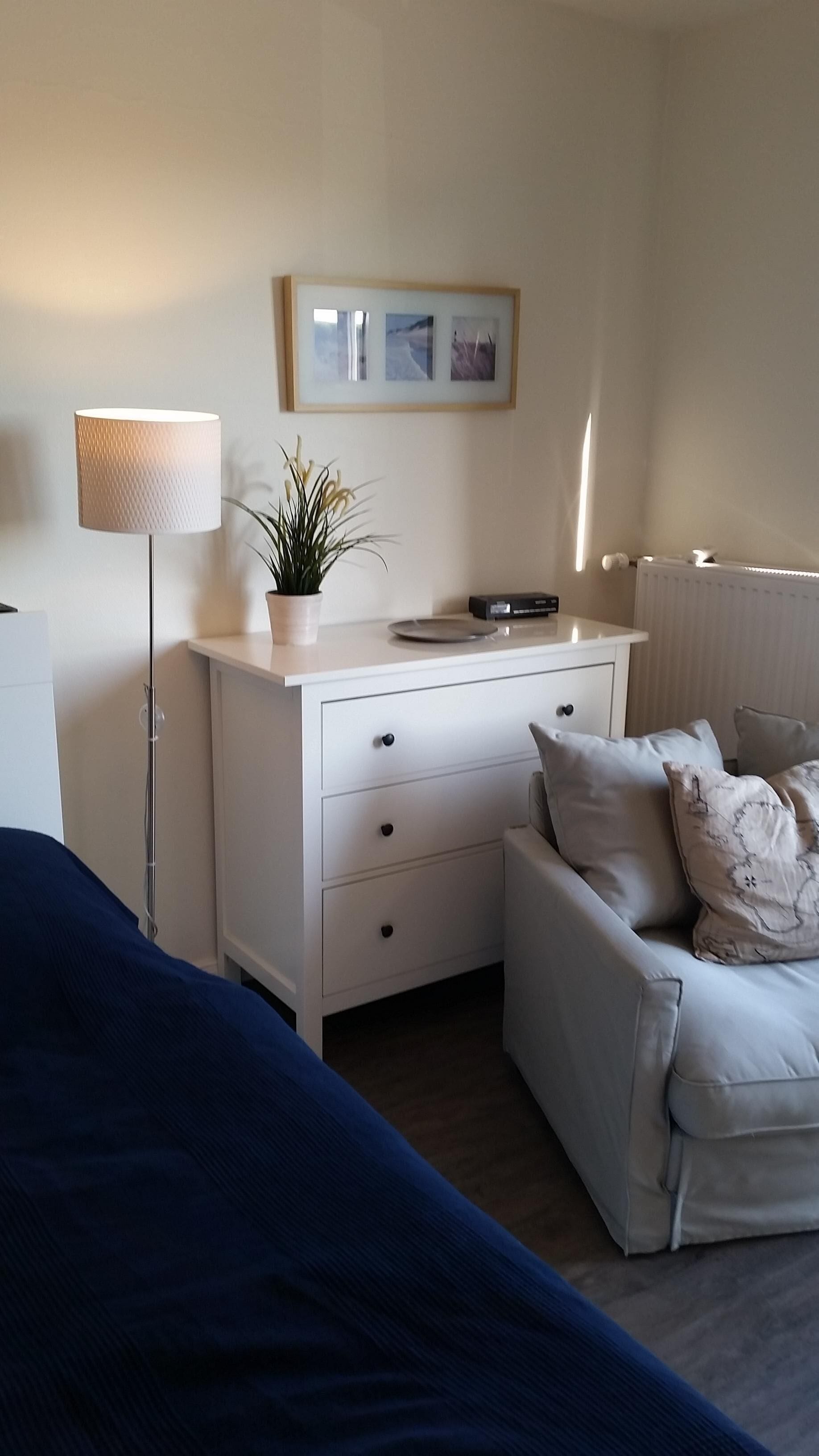 A bedroom with a bed, a bedside table with a lamp, a dresser, a radiator, and a wall-mounted picture.