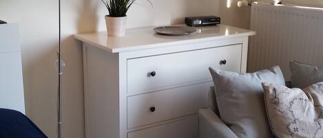 A bedroom with a bed, a bedside table with a lamp, a dresser, a radiator, and a wall-mounted picture.