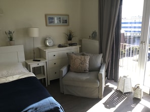 A hotel room with a bed, a desk, a chair, a clock, a plant, and a window with a view of buildings. - Newly renovated, close to the beach, sunny, south-facing balcony, WLAN (Sylt /Westerland)