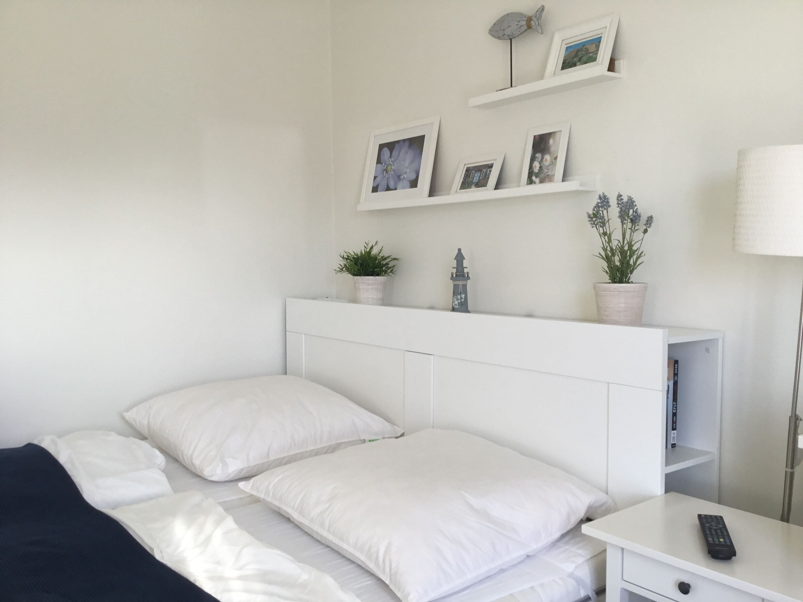 Une chambre à coucher avec un lit, des oreillers, une table de chevet, une lampe, une étagère avec des photos et des plantes, et une petite table avec une télécommande.