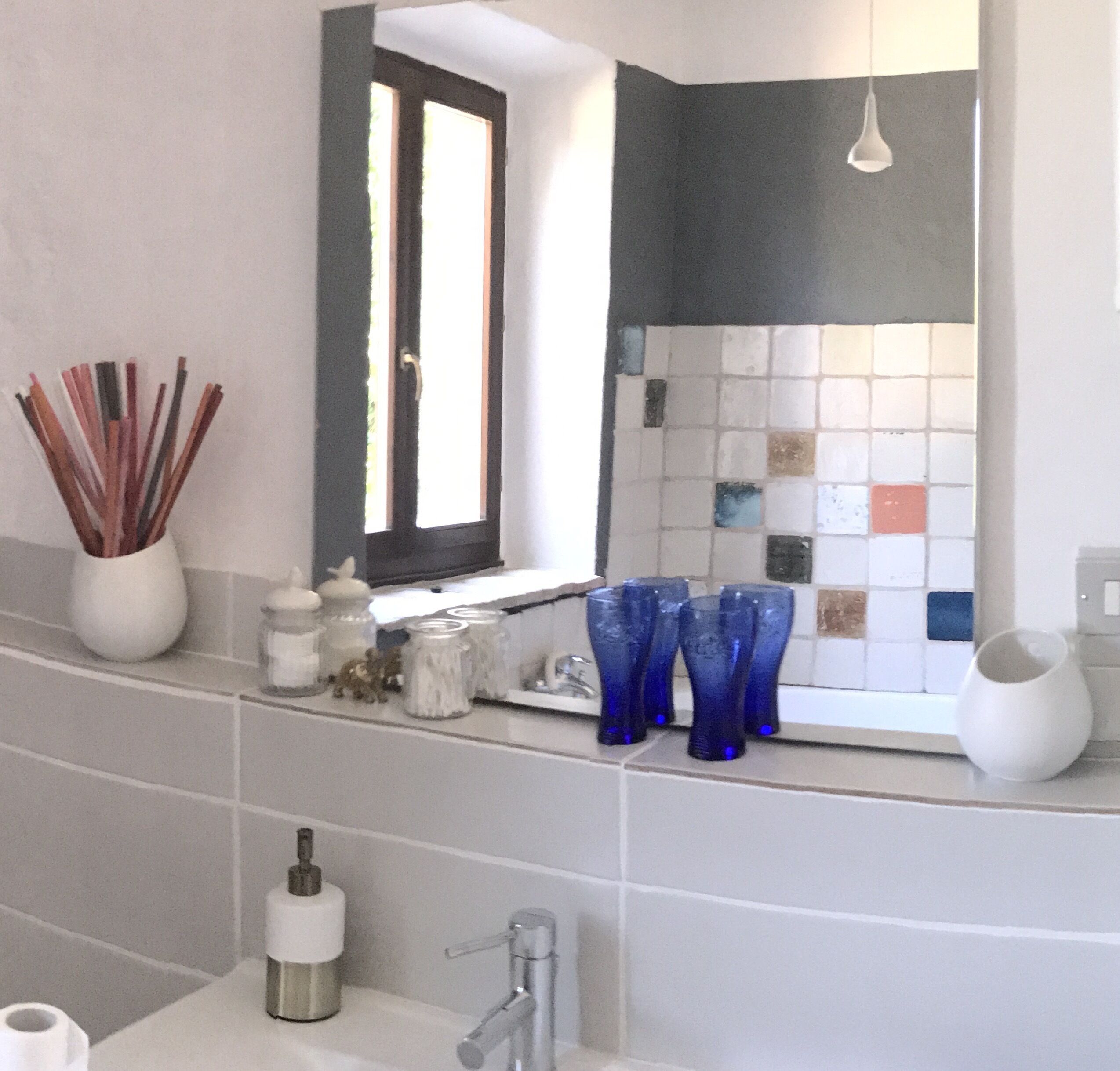 Un baño con un lavabo blanco, un espejo y una ventana. Hay jarrones de vidrio azul en la encimera.