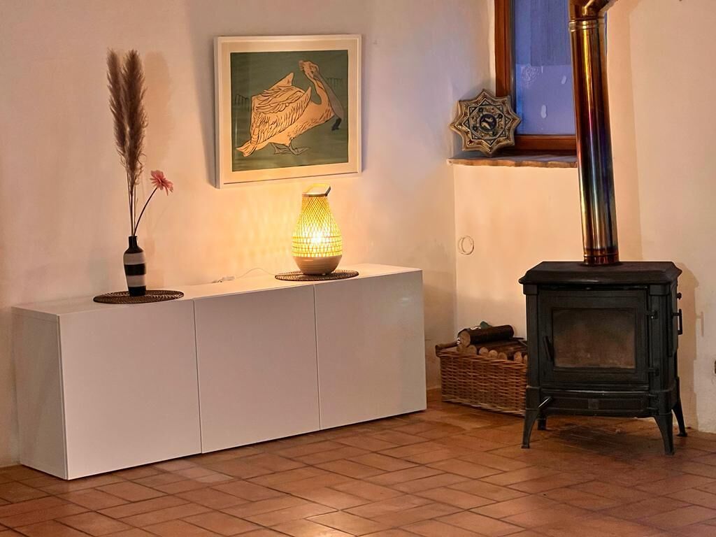 A room with a white console table, a decorative lamp, a framed artwork, a black stove, and a basket of logs.