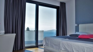 Ein modernes Schlafzimmer mit einem großen Bett, einem weißen Sofa und einer Glastür, die auf einen Balkon mit Meerblick führt.