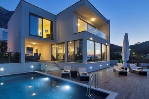Une maison moderne à deux étages, dotée d’une piscine, d’un espace salon extérieur et de grandes fenêtres.