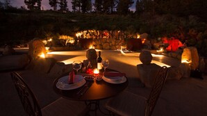 Un área de comedor al aire libre con paredes de piedra, un hogar de piedra y una mesa para dos personas con copas de vino, platos y velas.