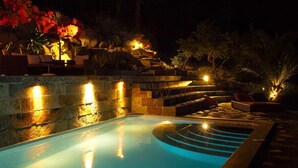 Una piscina nocturna con escalones de piedra y sillones de descanso, rodeada de vegetación ajardinada e iluminación cálida.