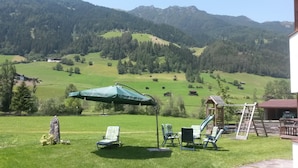 A grassy outdoor area with a swing set, a covered seating area with chairs, and a view of a mountainous landscape.