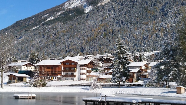 A snow-covered mountain village with wooden buildings, a lake, and a clear sky.