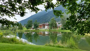 A lakeside view with a reflection of buildings, surrounded by greenery and mountains.