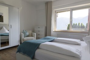 Un dormitorio con cama, armario, una silla y ventana con vistas a árboles y montañas.
