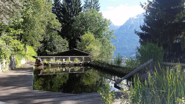 Eine Holzterrasse führt zu einem Pavillon über einem See, umgeben von dichtem Wald und Bergen.