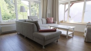 Ein modernes Wohnzimmer mit grauem Sofa, weißem Couchtisch und Blick ins Grüne durch große Fenster.