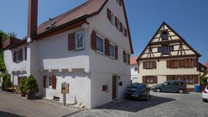 Une maison traditionnelle à colombages, dotée d’une cheminée, avec une voiture garée devant et un petit jardin agrémenté de plantes en pot.