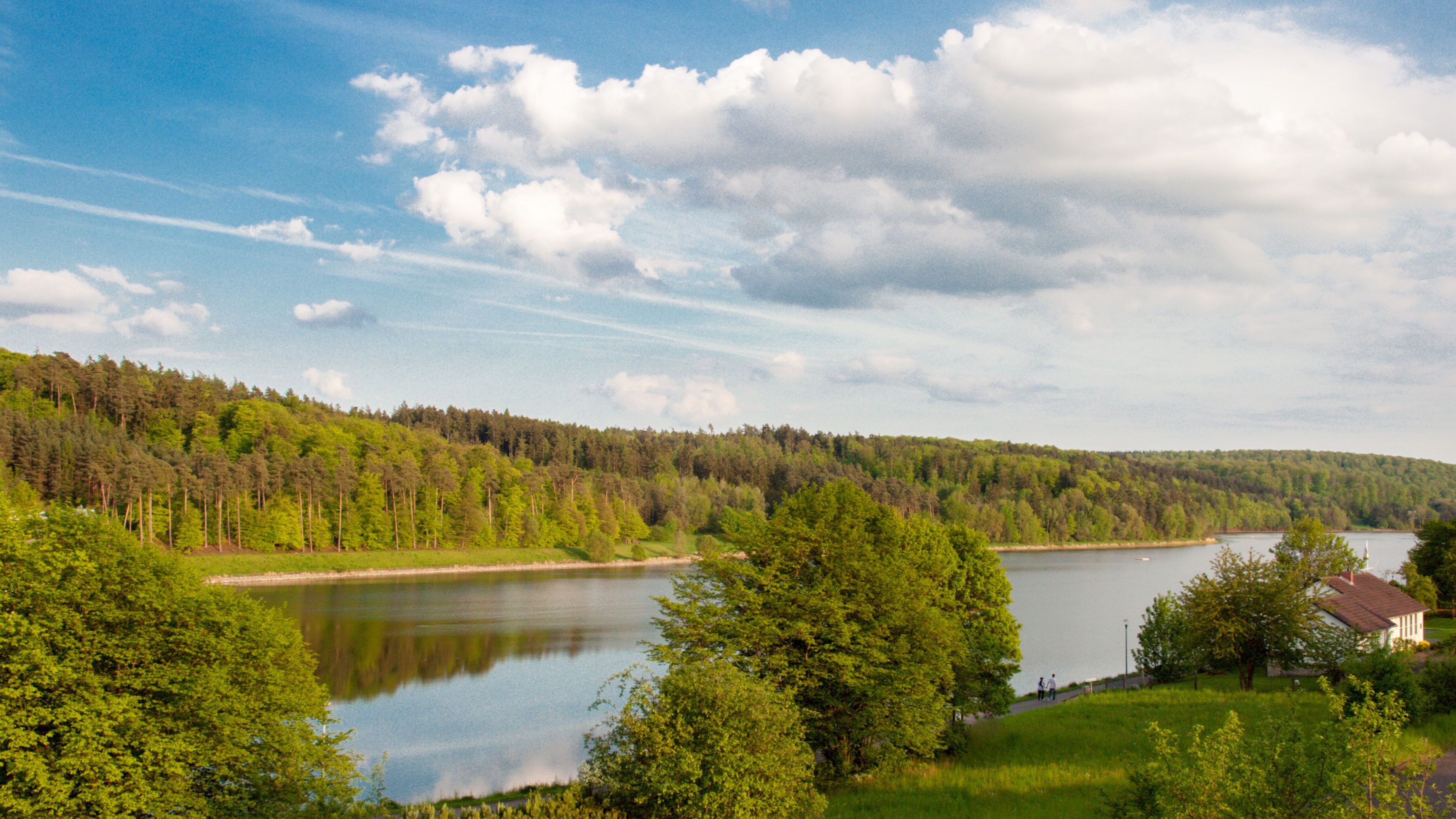 A lakeside view with a forested hill, a clear sky, and a small building near the water.