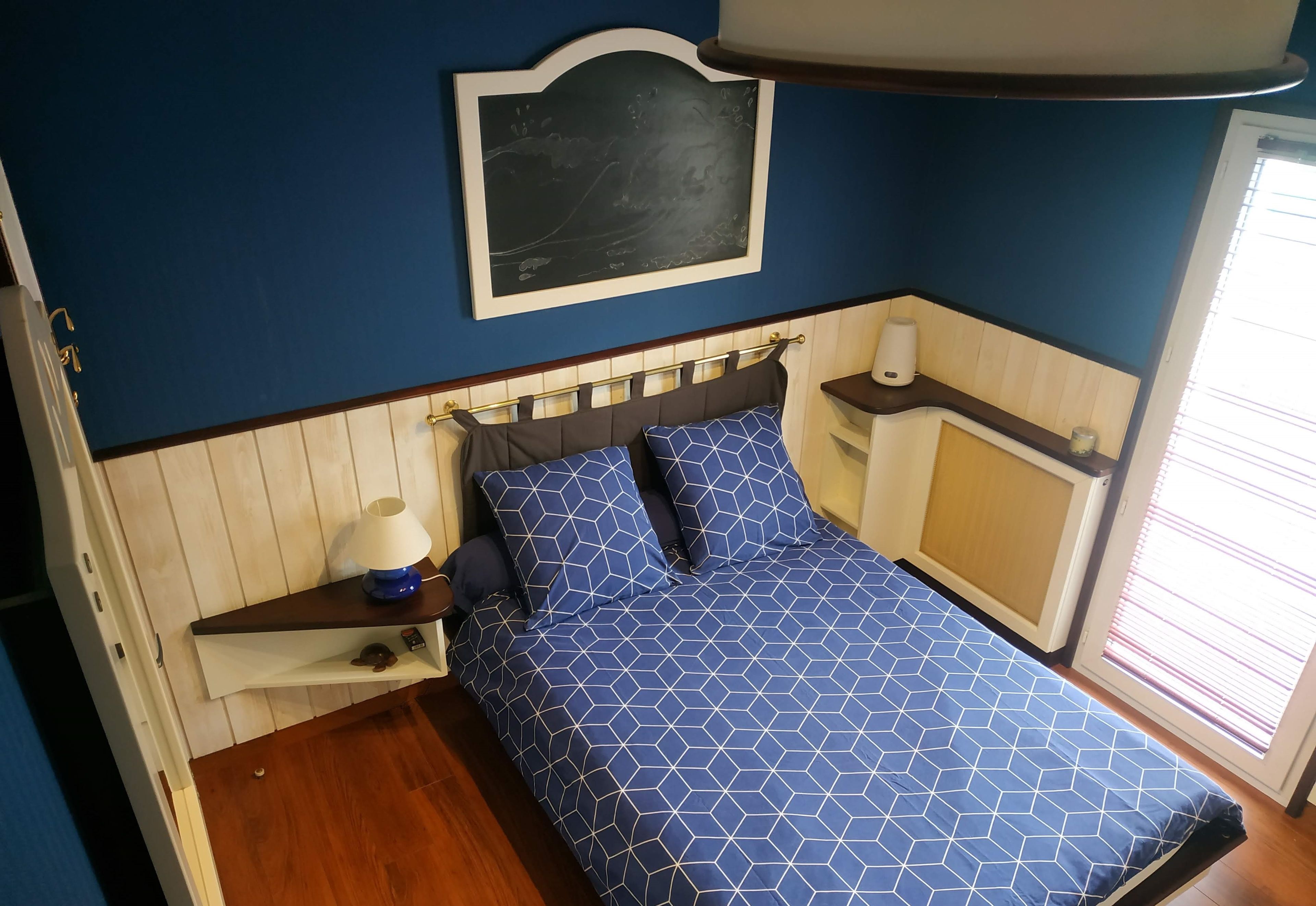 Une chambre à coucher comprenant un lit, une table de chevet, une étagère fixée au mur, une commode et une fenêtre avec des stores.