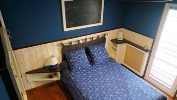 Une chambre à coucher comprenant un lit, une table de chevet, une étagère fixée au mur, une commode et une fenêtre avec des stores.