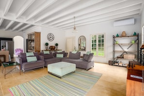 Una sala de estar amplia con un sofá modular grande, una mesa de centro y un tapete a rayas. Se observa un aparador de madera, una estantería y un gran ventanal que proporciona luz natural.
