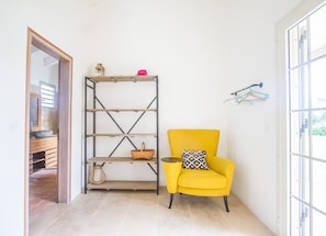 Une pièce avec un fauteuil jaune, une étagère en bois et une porte donnant sur une salle de bain.