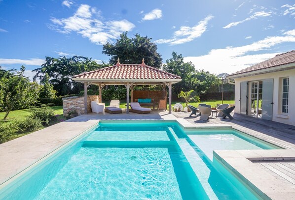 Una piscina con un pabellón y sillas de descanso, rodeada de un jardín ajardinado y una casa.