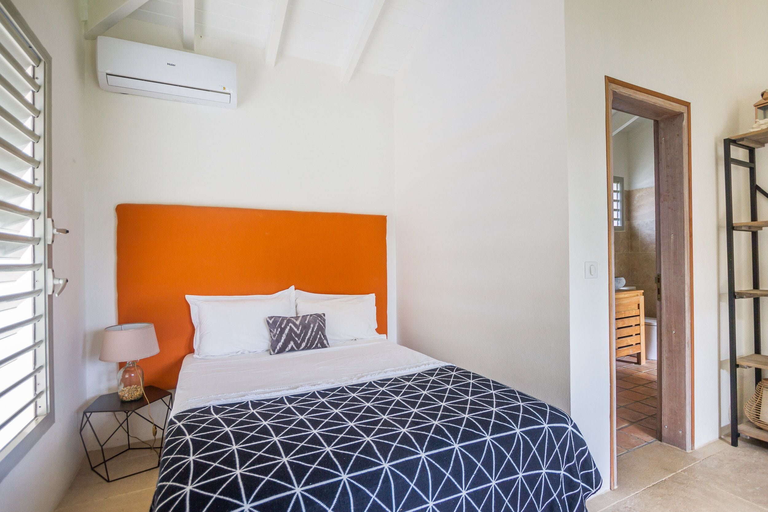 A hotel room with a bed, an air conditioner, a lamp, and a view of a wooden cabinet.