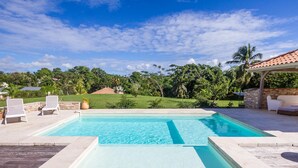 Una piscina rettangolare con superficie d'acqua cristallina azzurra, circondata da un solarium in pietra. Da un lato si vedono due poltrone bianche da relax e, dall'altro, una zona salotto coperta con un muro in pietra. La piscina è incorniciata da una vegetazione lussureggiante e un cielo azzurro terso.