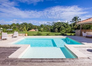 Ein rechteckiger Swimmingpool mit klar blauem Wasser, umgeben von einer Steindeck. Auf einer Seite befinden sich zwei weiße Liegestühle, auf der anderen eine überdachte Sitzgruppe mit einer Steinmauer. Der Pool wird von üppigem Grün und blauem Himmel begleitet.
