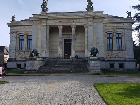 A classical building with a central entrance, flanked by statues and columns, and a gravel driveway leading to the entrance.