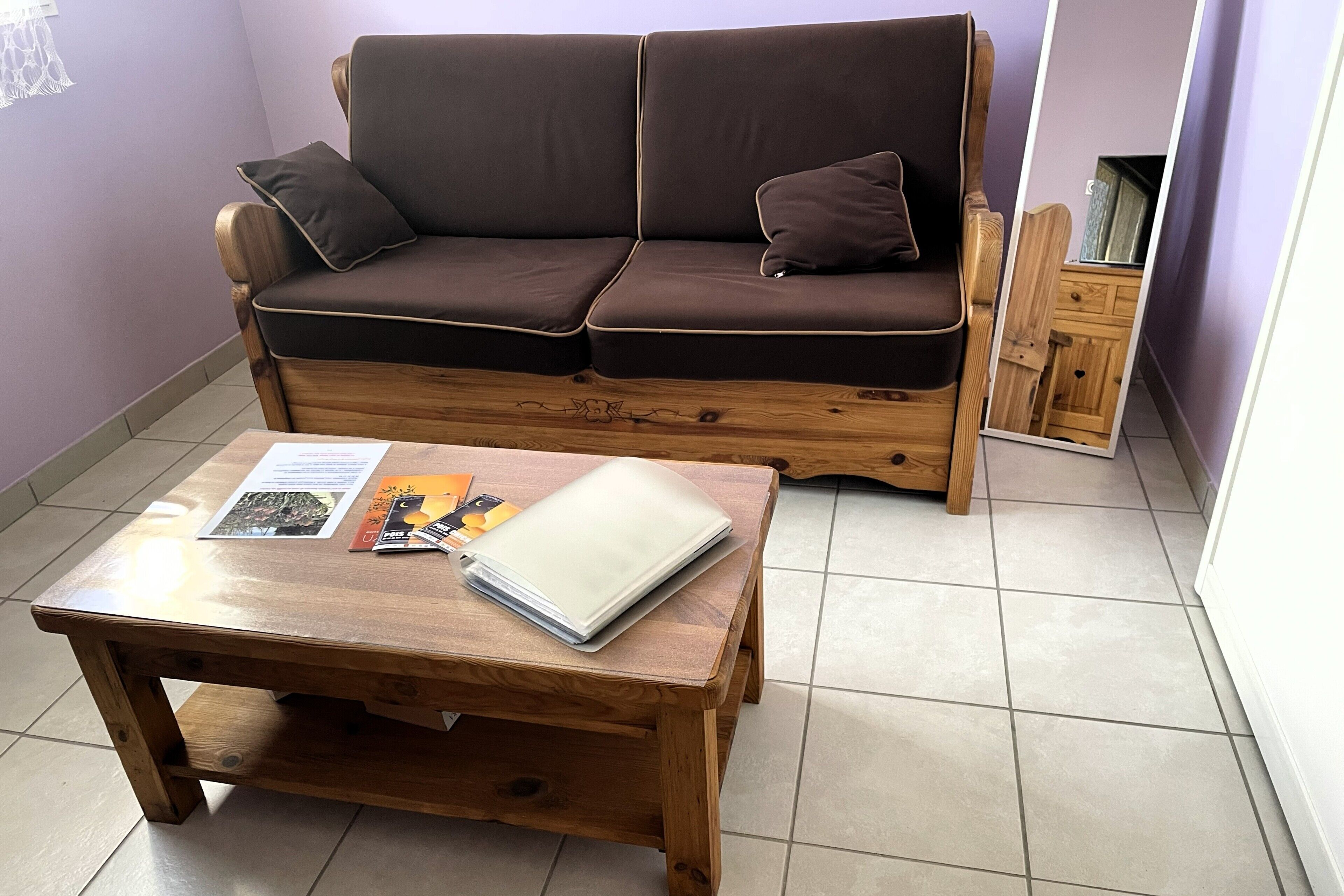 Un sofá de madera con cojines, una mesa de centro de madera con una revista y un espejo que refleja un aparador.