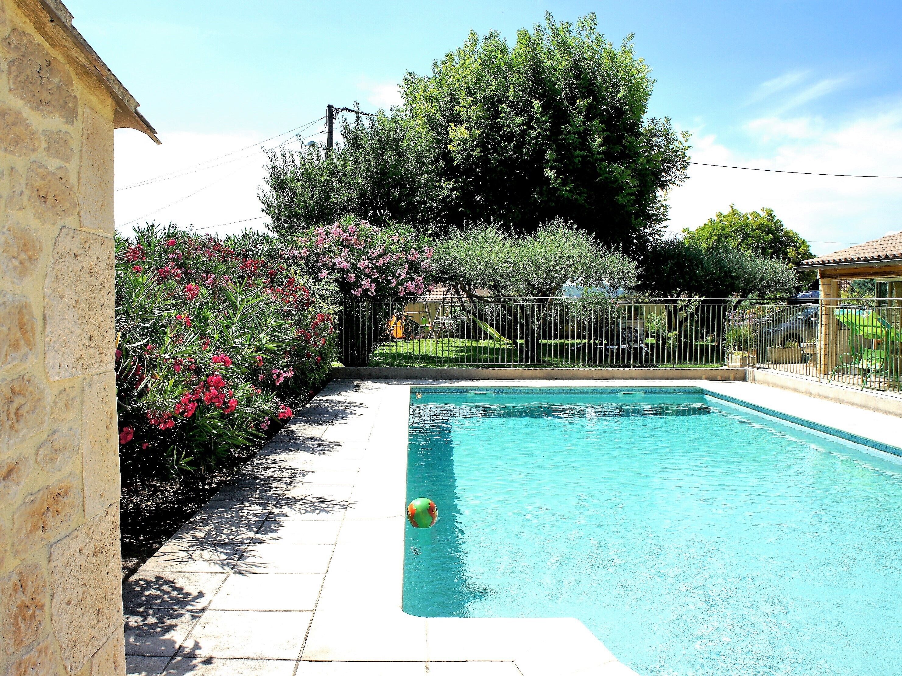 A swimming pool with a tiled deck, surrounded by a fence and lush greenery.