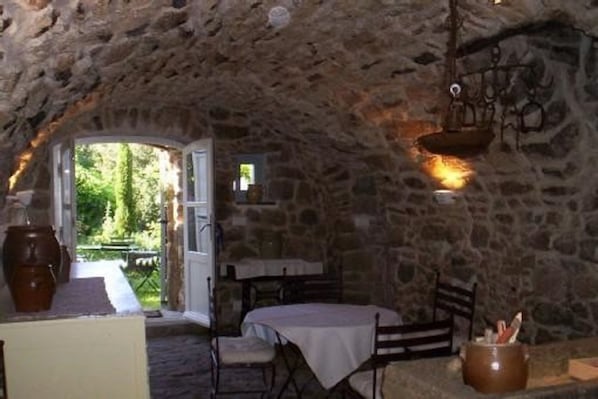 A rustic stone interior with a table and chairs, a hanging light fixture, and a view of greenery outside.