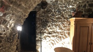 A stone-walled room with a wooden table and chairs, a wicker chair with a cushion, and a wooden cabinet.