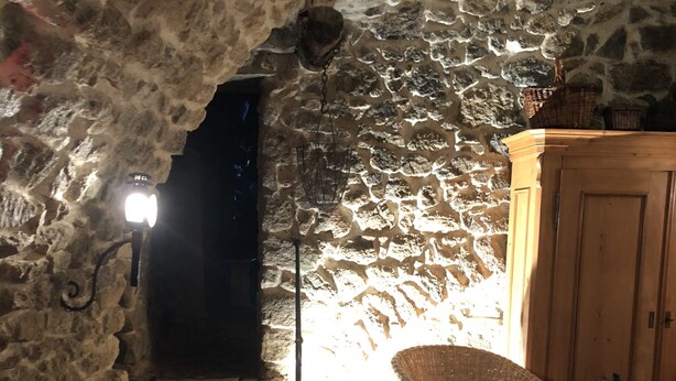 A stone-walled room with a wooden table and chairs, a wicker chair with a cushion, and a wooden cabinet. - Large independent apartment in a relaxing atmosphere overlooking the sea (Santa-Reparata-Di-Balagna)